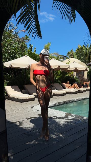 A woman in a red bikini and white cap dances playfully by a pool on a sunny day.