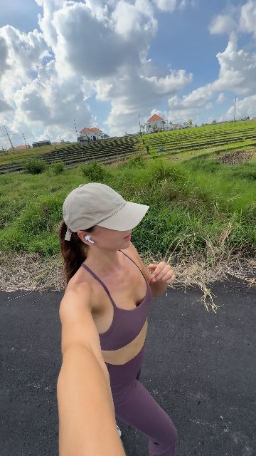A woman runs while holding a selfie camera on a path next to a green field.