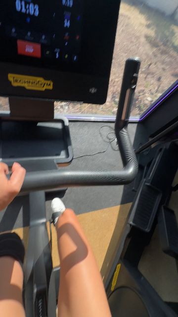 A woman uses a fitness machine and smiles at the camera in a gym setting.