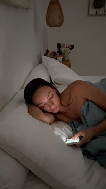 A woman lies in bed using her phone in a dimly lit room.