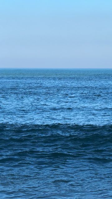 A high-angle shot captures waves consistently breaking on a sandy beach under a light blue sky.